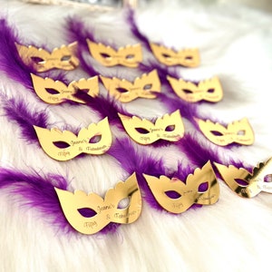 Puede incluir: M&aacute;scaras doradas de carnaval con plumas moradas. Las m&aacute;scaras est&aacute;n decoradas con el texto "Jeane's Fifty & Fabulous".