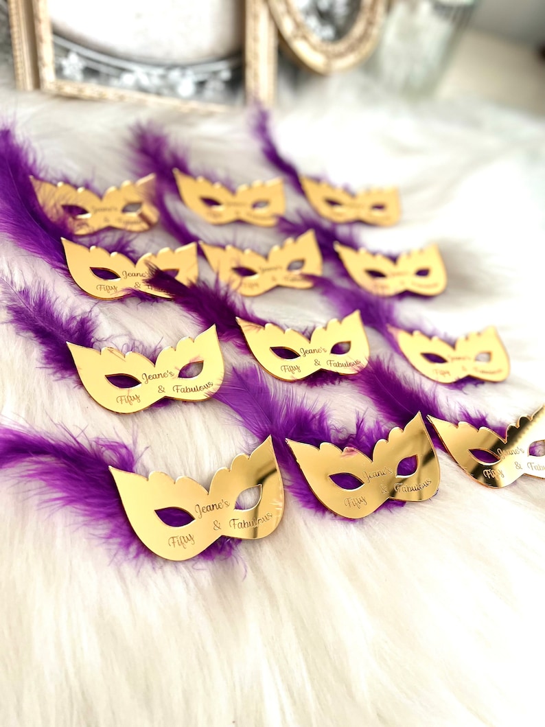 Puede incluir: M&aacute;scaras doradas de carnaval con plumas moradas. Las m&aacute;scaras est&aacute;n decoradas con el texto "Jeane's Fifty & Fabulous".