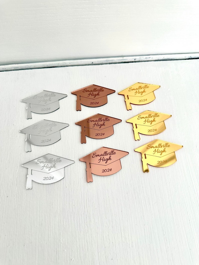 May include: Nine graduation caps made of mirrored acrylic in silver, gold, and rose gold. Each cap has the text "Smallville High 2024" engraved on it.