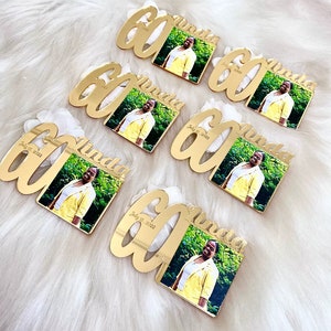 May include: Six gold-colored acrylic mirror plaques with the number 60 and the name "Linda" written in a cursive font. Each plaque has a small square photo of a woman in a yellow jacket. The date "July 15, 2022" is written on each plaque.