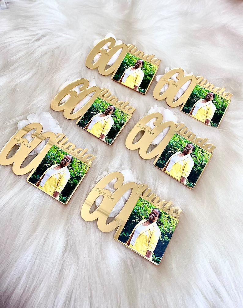 May include: Six gold-colored acrylic mirror plaques with the number 60 and the name "Linda" written in a cursive font. Each plaque has a small square photo of a woman in a yellow jacket. The date "July 15, 2022" is written on each plaque.