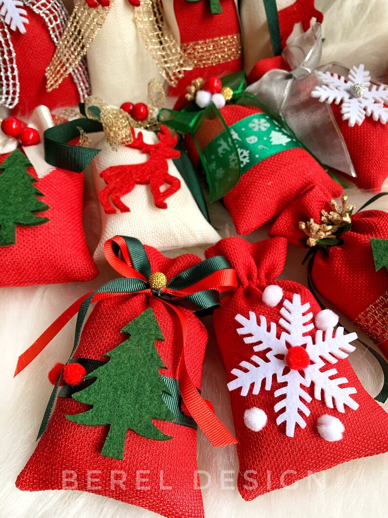 Puede incluir: Una colecci&oacute;n de peque&ntilde;as bolsas de regalo de tela roja con varias decoraciones con tem&aacute;tica navide&ntilde;a, que incluyen un reno, un copo de nieve, un &aacute;rbol de Navidad y una cinta. Las bolsas est&aacute;n llenas de peque&ntilde;os regalos y son perfectas para un calendario de adviento navide&ntilde;o.
