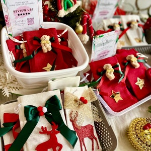 Puede incluir: Una cesta blanca llena de peque&ntilde;as bolsas de tela roja atadas con una cinta verde y decoradas con un peque&ntilde;o osito de peluche. Las bolsas est&aacute;n sobre una mesa con un cartel que dice "Hello, Thank you for being a part of our family, REVIEW US!" con cinco estrellas. Hay otras cestas llenas de bolsas similares en el fondo.