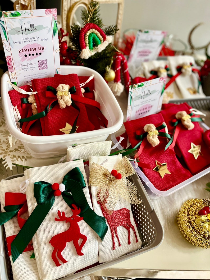 Puede incluir: Una cesta blanca llena de peque&ntilde;as bolsas de tela roja atadas con una cinta verde y decoradas con un peque&ntilde;o osito de peluche. Las bolsas est&aacute;n sobre una mesa con un cartel que dice "Hello, Thank you for being a part of our family, REVIEW US!" con cinco estrellas. Hay otras cestas llenas de bolsas similares en el fondo.