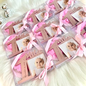 May include: Pink and gold favor boxes with a clear lid, each containing a pink flower, a rose gold number one, and a photo of a baby. The boxes are tied with a pink ribbon and have the name "Mila" and the date "November 12, 2021" printed on them.