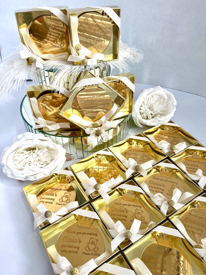May include: Gold favor boxes with a round mirror inside. Each mirror has a personalized message with a teddy bear graphic and the text "Thank you for making Olivia's first birthday very special".