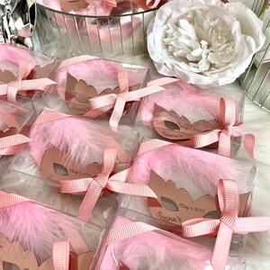 May include: Pink and gold party favours in clear boxes with pink feathers and a pink ribbon. Each box has a gold mirror mask with the text "Sienna's" and the date "May 6, 2023".