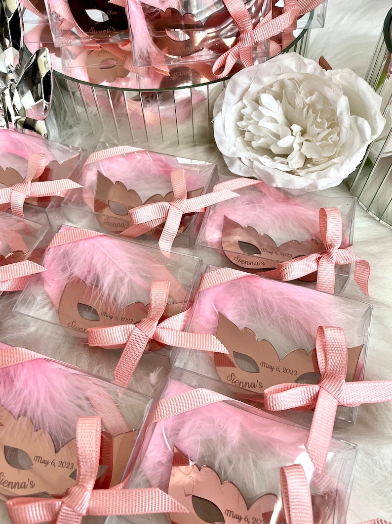 May include: Pink and gold party favours in clear boxes with pink feathers and a pink ribbon. Each box has a gold mirror mask with the text "Sienna's" and the date "May 6, 2023".