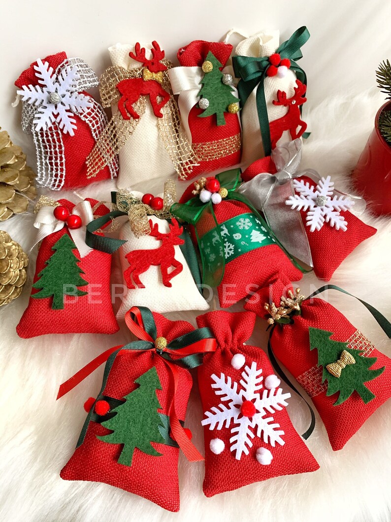 Puede incluir: Surtido de bolsas de regalo de tela de Navidad rojas y blancas con varios adornos de fieltro, incluyendo copos de nieve, renos y &aacute;rboles de Navidad. Las bolsas est&aacute;n atadas con cinta verde, roja y dorada.