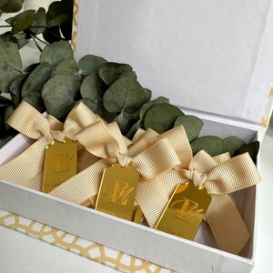 May include: Three gold gift tags with a bow tied around each one. The tags are rectangular and have the letter "E" engraved on them. The tags are in a white box with a gold pattern on the bottom.