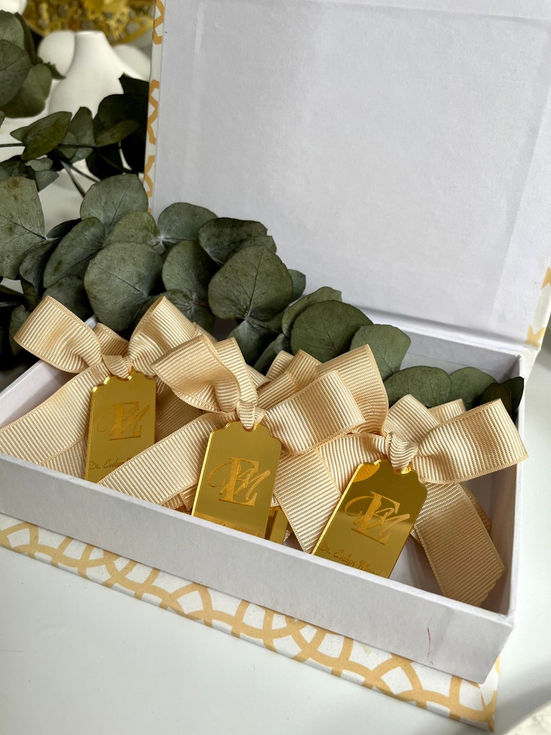 May include: Three gold gift tags with a bow tied around each one. The tags are rectangular and have the letter "E" engraved on them. The tags are in a white box with a gold pattern on the bottom.