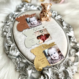May include: Gold, silver, and rose gold bear-shaped photo frames with a baby's photo and the baby's name and birthdate. The frames are on a silver tray with an ornate design.