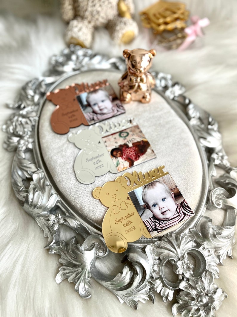 May include: Gold, silver, and rose gold bear-shaped photo frames with a baby's photo and the baby's name and birthdate. The frames are on a silver tray with an ornate design.