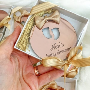 Op de afbeelding: Een roze gouden spiegel met een uitsparing van twee babyvoetjes en de tekst "Nan's baby shower" in een witte doos met een gouden lint en strik.
