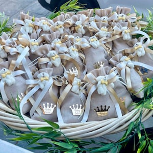 May include: A basket filled with small beige fabric bags tied with white and gold ribbon. Each bag has a small white teddy bear with a gold bow tie attached to it. The bags have gold crown stickers with the text "Baby Sela" on them.