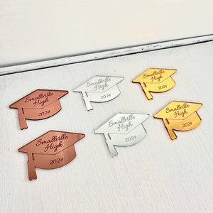 May include: Five graduation cap-shaped mirror ornaments in different colors: silver, gold, rose gold, copper, and black. Each ornament has the text "Smallville High 2024" engraved on it.