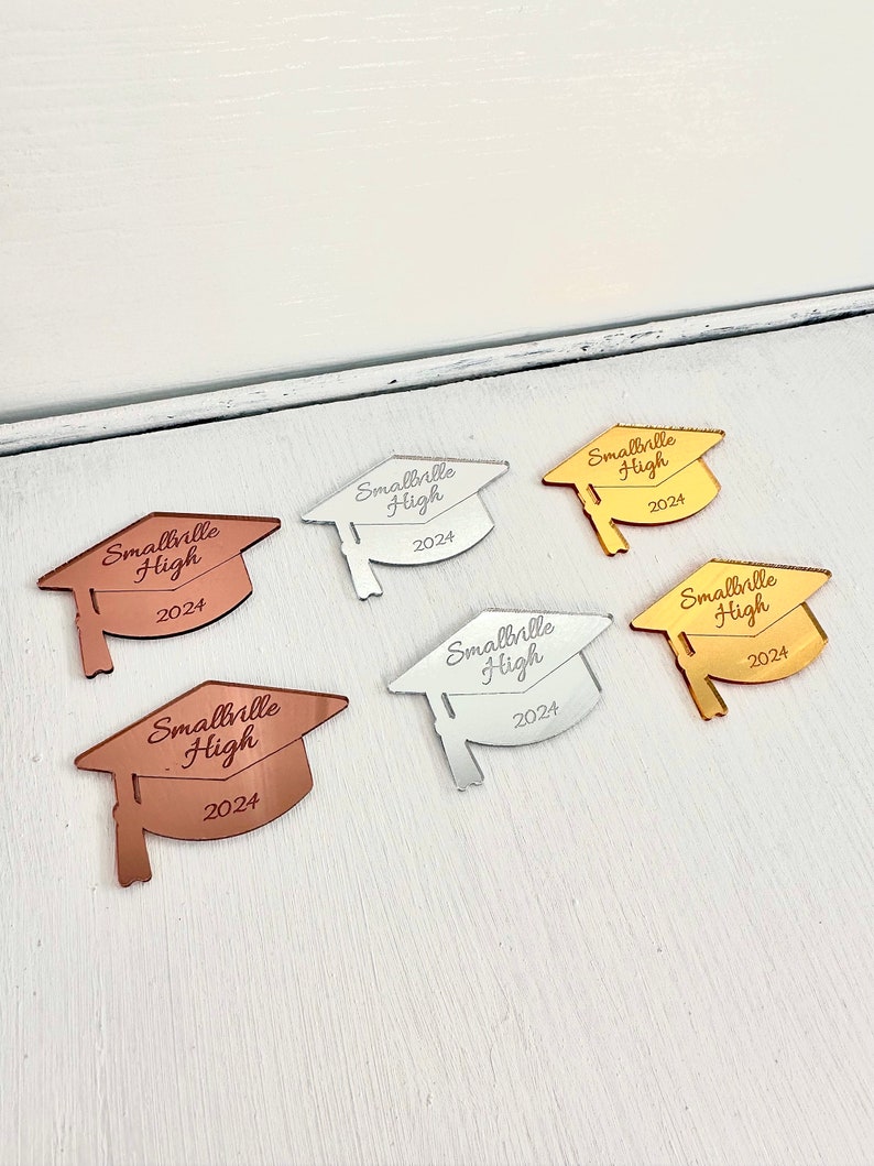 May include: Five graduation cap-shaped mirror ornaments in different colors: silver, gold, rose gold, copper, and black. Each ornament has the text "Smallville High 2024" engraved on it.