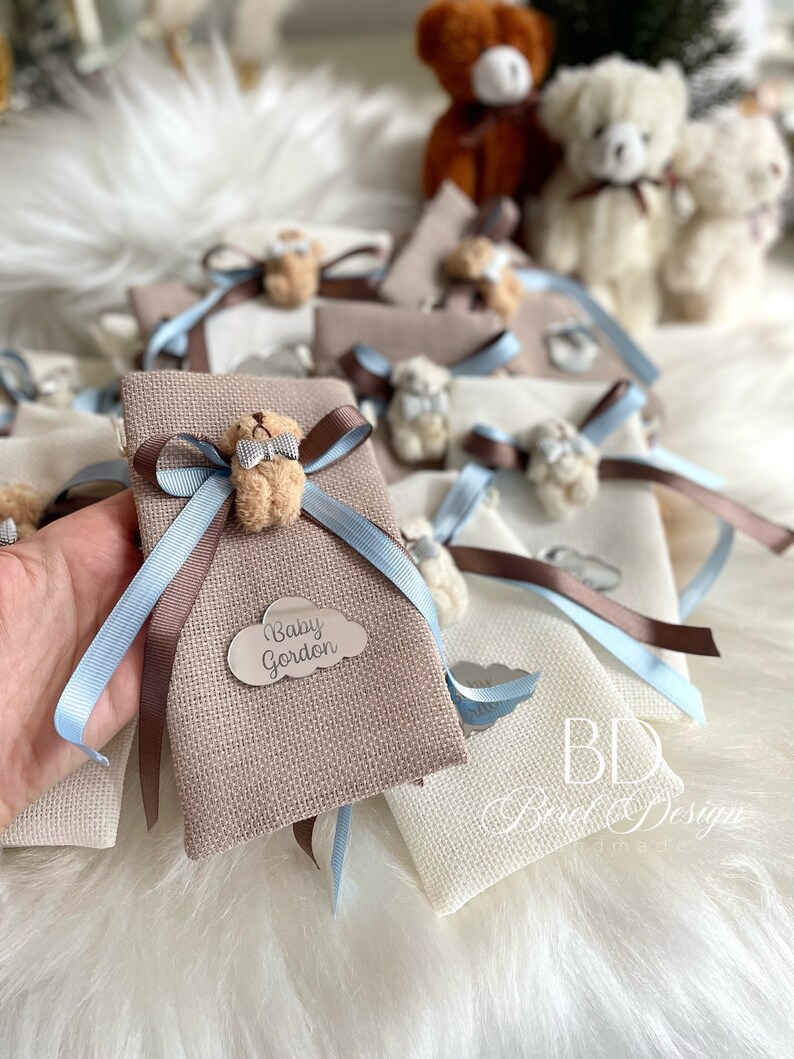 May include: A collection of small, brown fabric gift bags with blue and brown ribbon. Each bag has a small, brown teddy bear with a bow tie and a silver tag with the text "Baby Gordon".