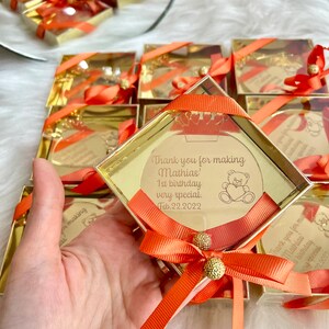 May include: Gold favor boxes with orange ribbon bows and a clear acrylic circle with a personalized message that says "Thank you for making Mathias' 1st birthday very special. Feb. 22. 2022". The circle also has a small cartoon bear with a heart.