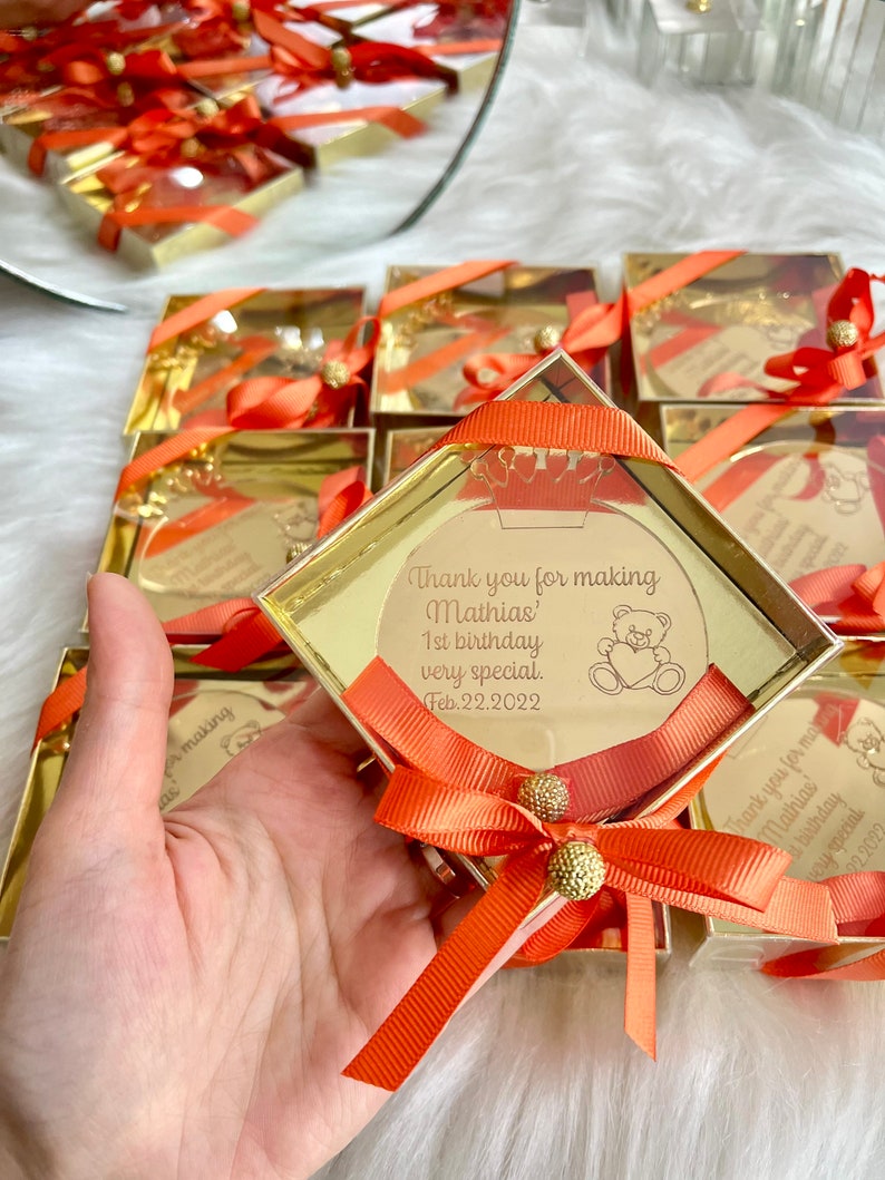 May include: Gold favor boxes with orange ribbon bows and a clear acrylic circle with a personalized message that says "Thank you for making Mathias' 1st birthday very special. Feb. 22. 2022". The circle also has a small cartoon bear with a heart.