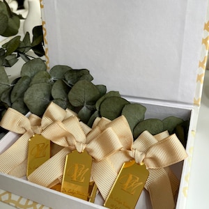 May include: A white box with a gold patterned design. Inside the box are four gold mirror-like tags with a cursive letter "E" and the text "Dr. Emily Maxwell 05/15/2022". Each tag is tied with a beige satin ribbon.