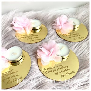 May include: Gold mirrored round candle holders with a pink flower on top and a small white tea light candle. The holders have a personalized message engraved on them that says "I'm lit and so are you! Thank you for celebrating my 30th with me. Love, Rochelle"