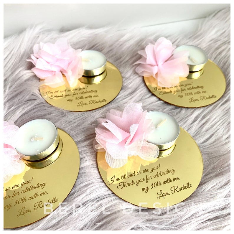 May include: Gold mirrored round candle holders with a pink flower on top and a small white tea light candle. The holders have a personalized message engraved on them that says "I'm lit and so are you! Thank you for celebrating my 30th with me. Love, Rochelle"