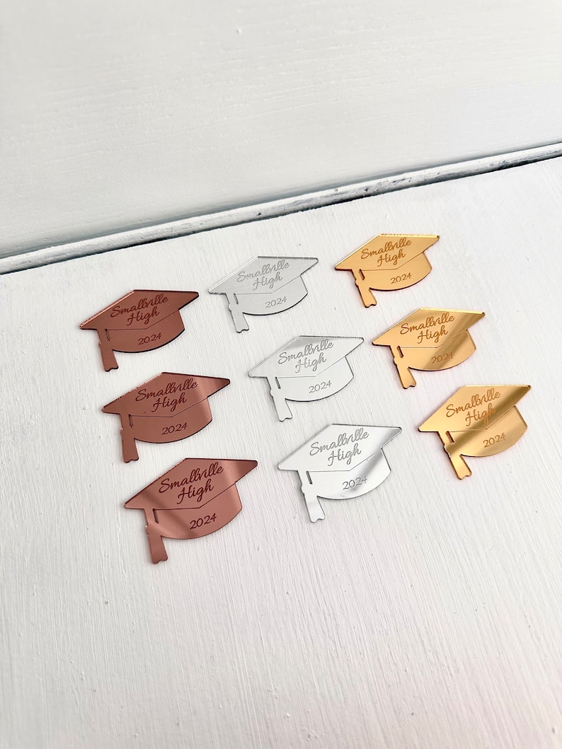 May include: Eight graduation cap-shaped mirror ornaments in gold, silver, and rose gold. Each ornament features the text "Smallville High 2024".