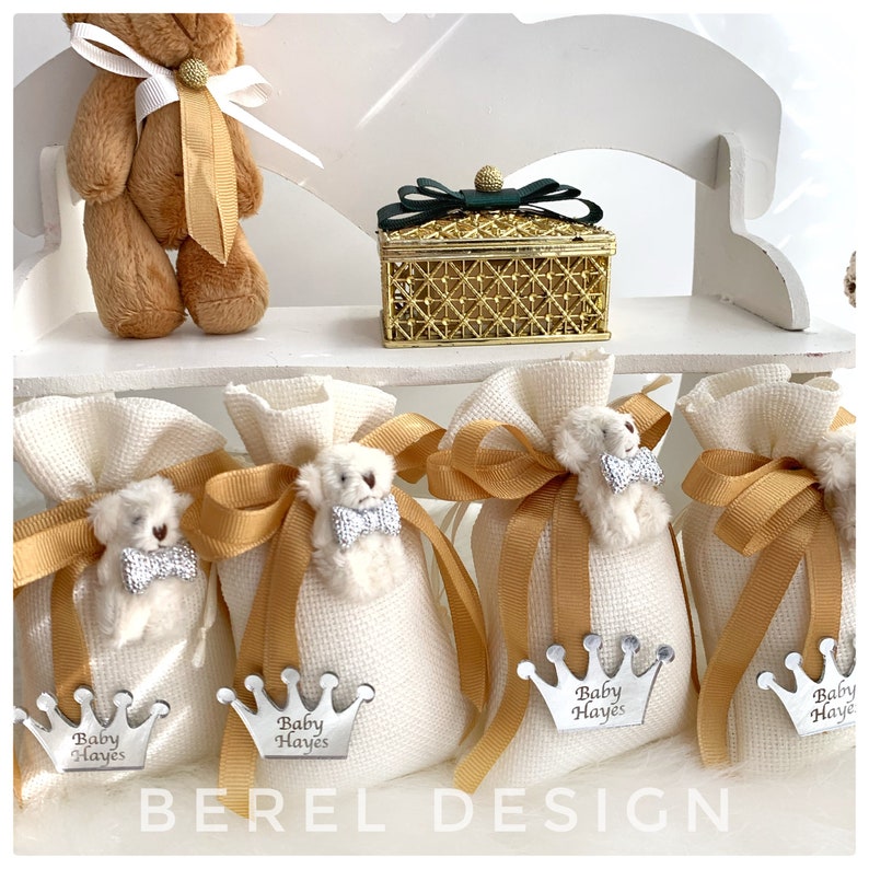May include: Four white favor bags with gold ribbon and a small teddy bear with a bow tie. Each bag has a silver crown with the text "Baby Hayes" on it. There is a gold box with a green bow on the top shelf.