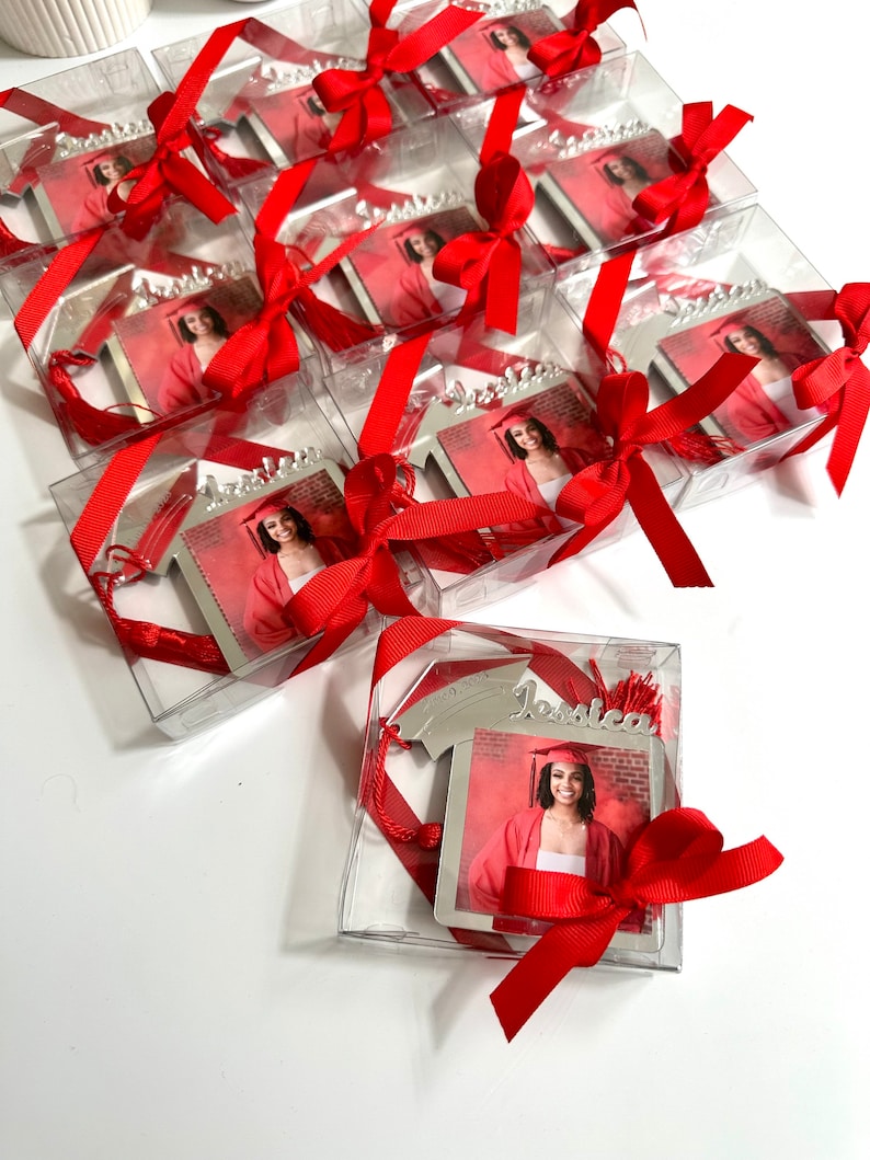 May include: Silver graduation bookmark with a photo frame, red tassel, and red ribbon. The bookmark is in a clear plastic box with a red ribbon bow. The bookmark has the name 'Jessica' engraved on it.