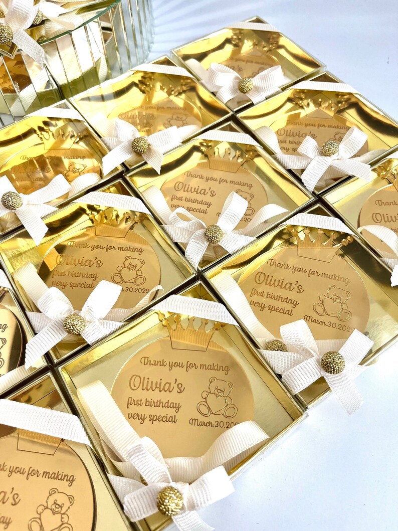 May include: Gold favor boxes with a round, gold mirror with a teddy bear and the text "Thank you for making Olivia's first birthday very special March 30, 2023".