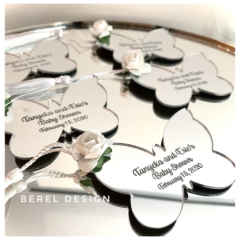 Puede incluir: Favores de espejo en forma de mariposa plateados con un adorno de flor blanca y un mensaje personalizado que dice "Tanyeka and Eric's Baby Shower February 15, 2020".