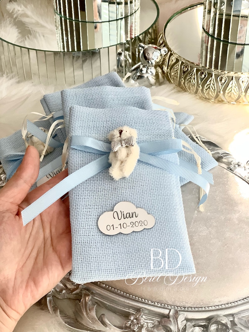 Puede incluir: Bolsas de regalo de tela azul claro con un osito de peluche blanco y una etiqueta en forma de nube blanca con el nombre "Vian" y la fecha "01-10-2020".
