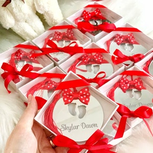 Op de afbeelding: Een set kleine, witte geschenkdozen met rood lint en een rode strik. Elke doos bevat een ronde, zilveren spiegel met een uitsnijding van babyvoetjes en de naam "Skylar Dawn" erop gegraveerd.