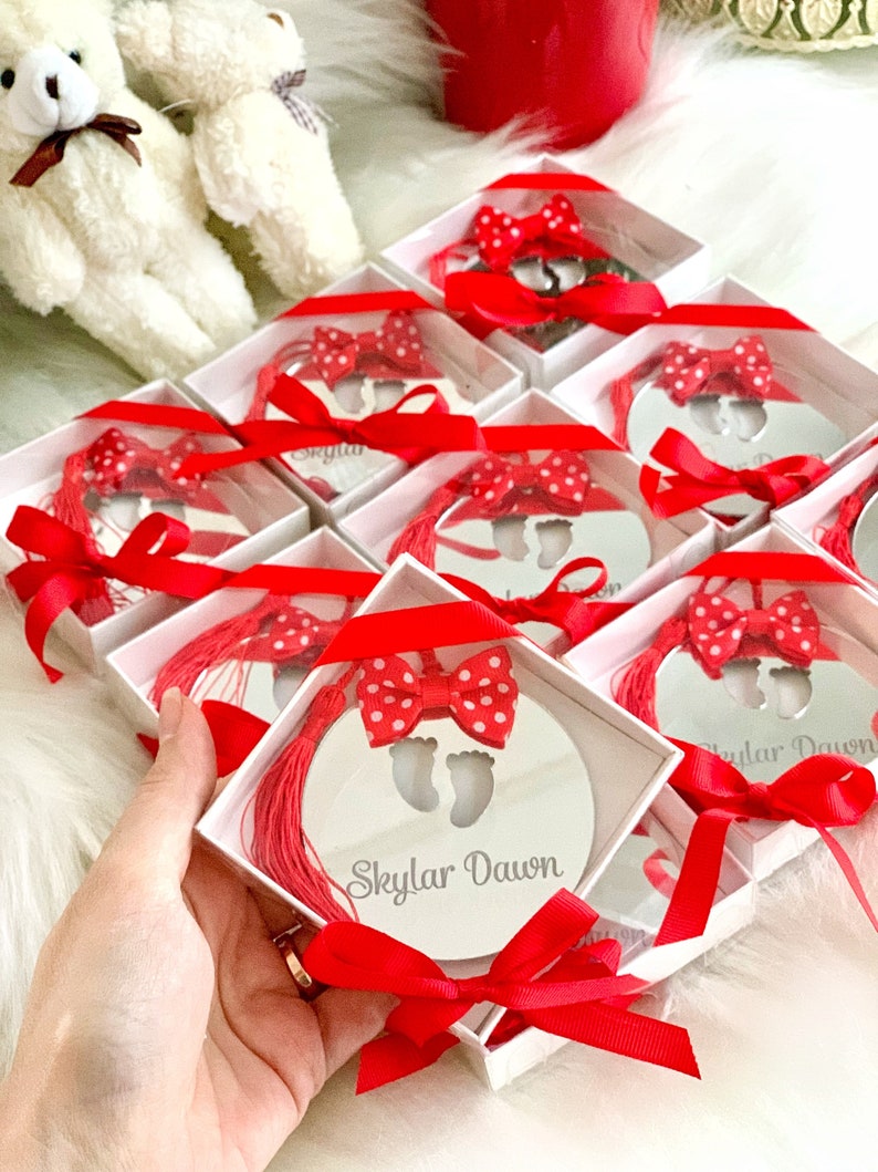Op de afbeelding: Een set kleine, witte geschenkdozen met rood lint en een rode strik. Elke doos bevat een ronde, zilveren spiegel met een uitsnijding van babyvoetjes en de naam "Skylar Dawn" erop gegraveerd.