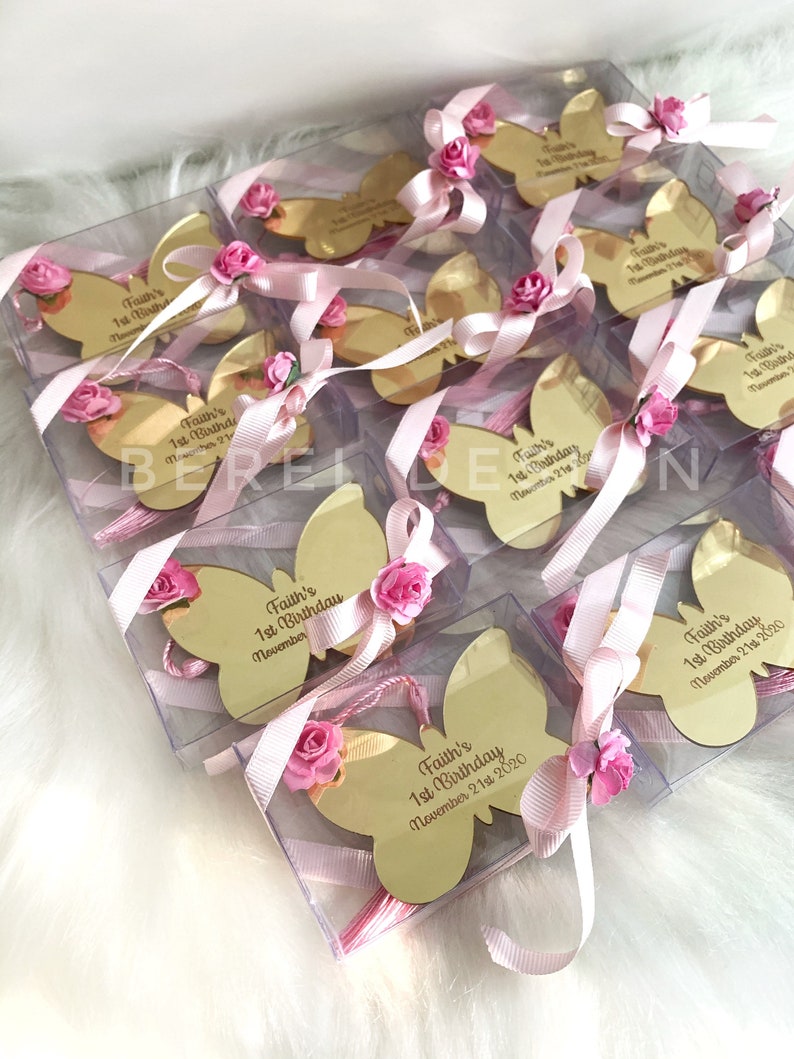 Puede incluir: Favores de espejo en forma de mariposa dorada con cinta rosa y flores. Cada espejo tiene el texto "Faith's 1st Birthday" y la fecha "November 21st 2020".
