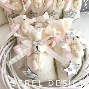 May include: White fabric gift bags with pink ribbon bows and a small white teddy bear with a silver bow. Each bag has a silver star with the name "Sofia" on it.
