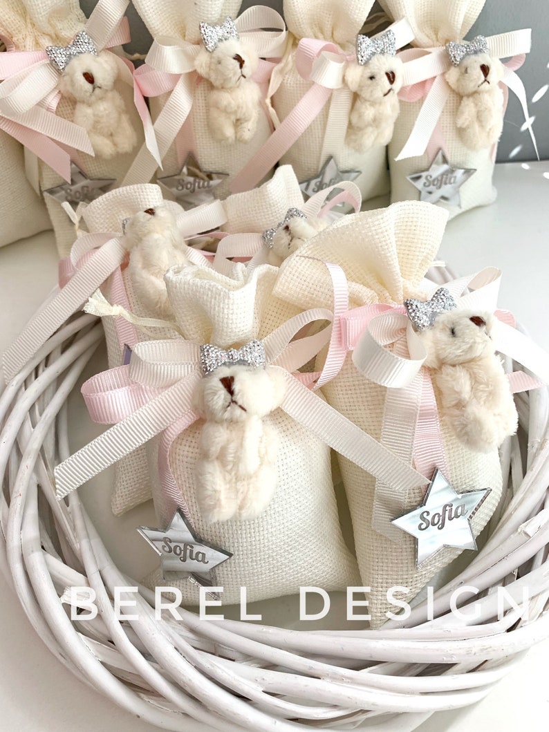 May include: White fabric gift bags with pink ribbon bows and a small white teddy bear with a silver bow. Each bag has a silver star with the name "Sofia" on it.