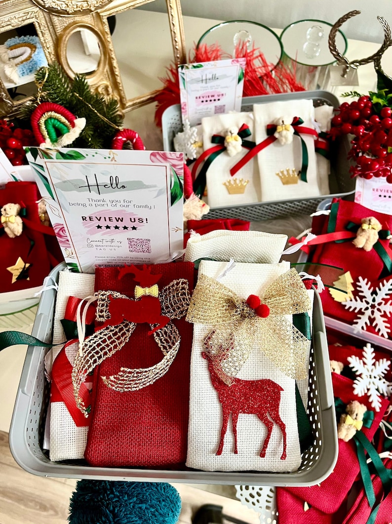 Puede incluir: Una cesta gris llena de peque&ntilde;as bolsas de regalo rojas y blancas. Las bolsas est&aacute;n decoradas con lazos, renos y estrellas. Un cartel en el fondo dice "Hola, gracias por ser parte de nuestra familia! &iexcl;Calif&iacute;canos!" con cinco estrellas.