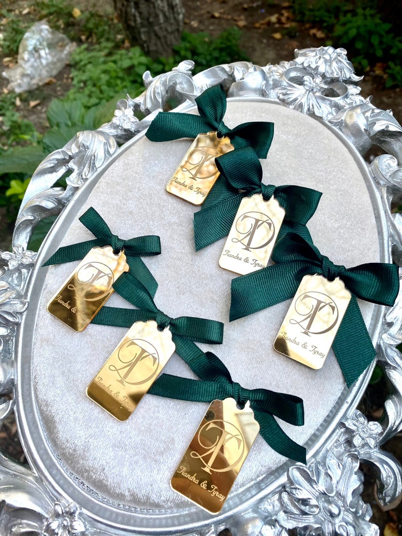 May include: Five gold-colored rectangular tags with a decorative border and the text "Tiandra & Tyray" and a letter "T" in a decorative font. Each tag is tied with a dark green satin ribbon bow.