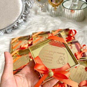 May include: A small, clear box with a gold lid contains a round, gold mirror with a thank you message for making Mathias' 1st birthday very special. The message is dated Feb. 22, 2022. The box is tied with a bright orange ribbon and a gold bow.