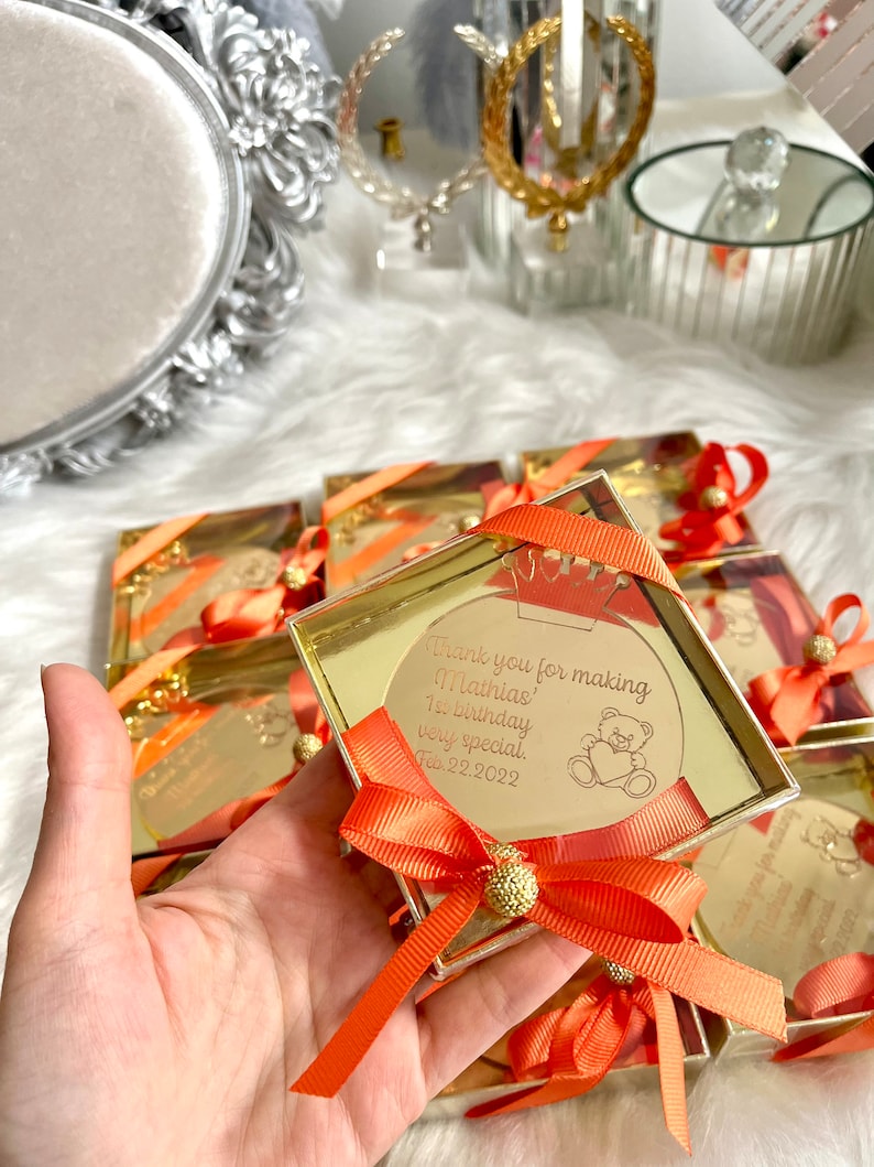 May include: A small, clear box with a gold lid contains a round, gold mirror with a thank you message for making Mathias' 1st birthday very special. The message is dated Feb. 22, 2022. The box is tied with a bright orange ribbon and a gold bow.