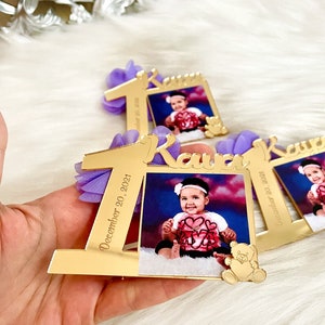 Puede incluir: Marco de fotos de acr&iacute;lico espejo dorado con un lazo morado y el n&uacute;mero uno, el nombre "Kava" y la fecha "20 de diciembre de 2021". Una foto de una ni&ntilde;a peque&ntilde;a est&aacute; en el marco.