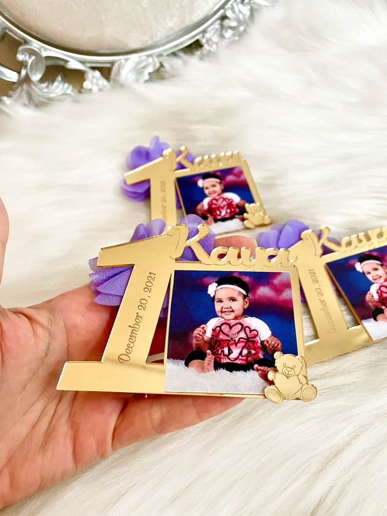 Puede incluir: Marco de fotos de acr&iacute;lico espejo dorado con un lazo morado y el n&uacute;mero uno, el nombre "Kava" y la fecha "20 de diciembre de 2021". Una foto de una ni&ntilde;a peque&ntilde;a est&aacute; en el marco.