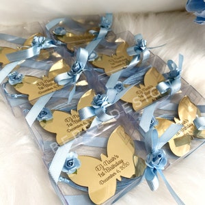 Puede incluir: Favores de espejo en forma de mariposa dorada con cinta azul y detalles de flores. Cada espejo presenta un mensaje personalizado con un nombre y el texto "1er cumplea&ntilde;os" y una fecha.