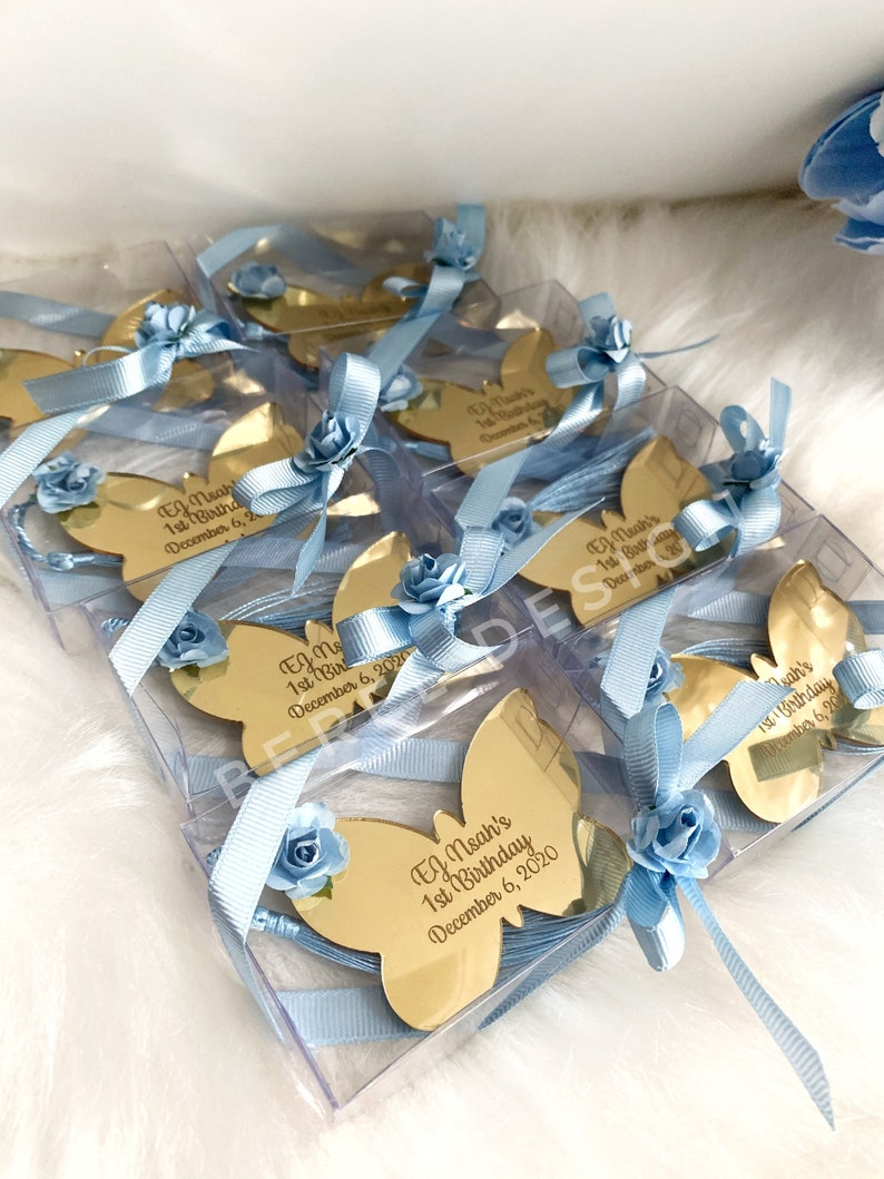 Puede incluir: Favores de espejo en forma de mariposa dorada con cinta azul y detalles de flores. Cada espejo presenta un mensaje personalizado con un nombre y el texto "1er cumplea&ntilde;os" y una fecha.