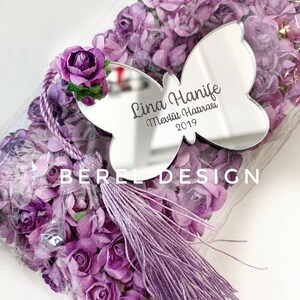 Puede incluir: Un espejo en forma de mariposa p&uacute;rpura con el texto "Lina Hanife Mevlit Hatırası 2019" grabado en &eacute;l. El espejo est&aacute; rodeado de flores de papel p&uacute;rpura y una borla p&uacute;rpura.