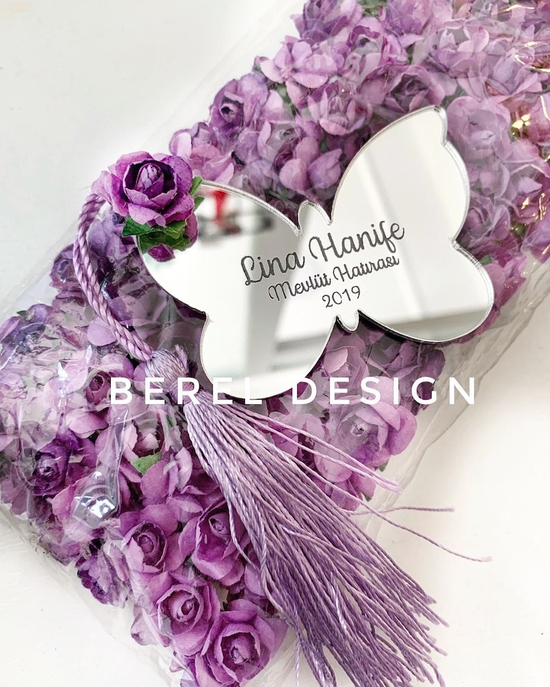 Puede incluir: Un espejo en forma de mariposa p&uacute;rpura con el texto "Lina Hanife Mevlit Hatırası 2019" grabado en &eacute;l. El espejo est&aacute; rodeado de flores de papel p&uacute;rpura y una borla p&uacute;rpura.