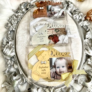 May include: Three gold-colored teddy bear shaped photo frames with a picture of a baby boy. The frames are in clear plastic boxes with gold and silver ribbons. The frames have the baby's name "Oliver" and the date "September 24th, 2022" engraved on them.