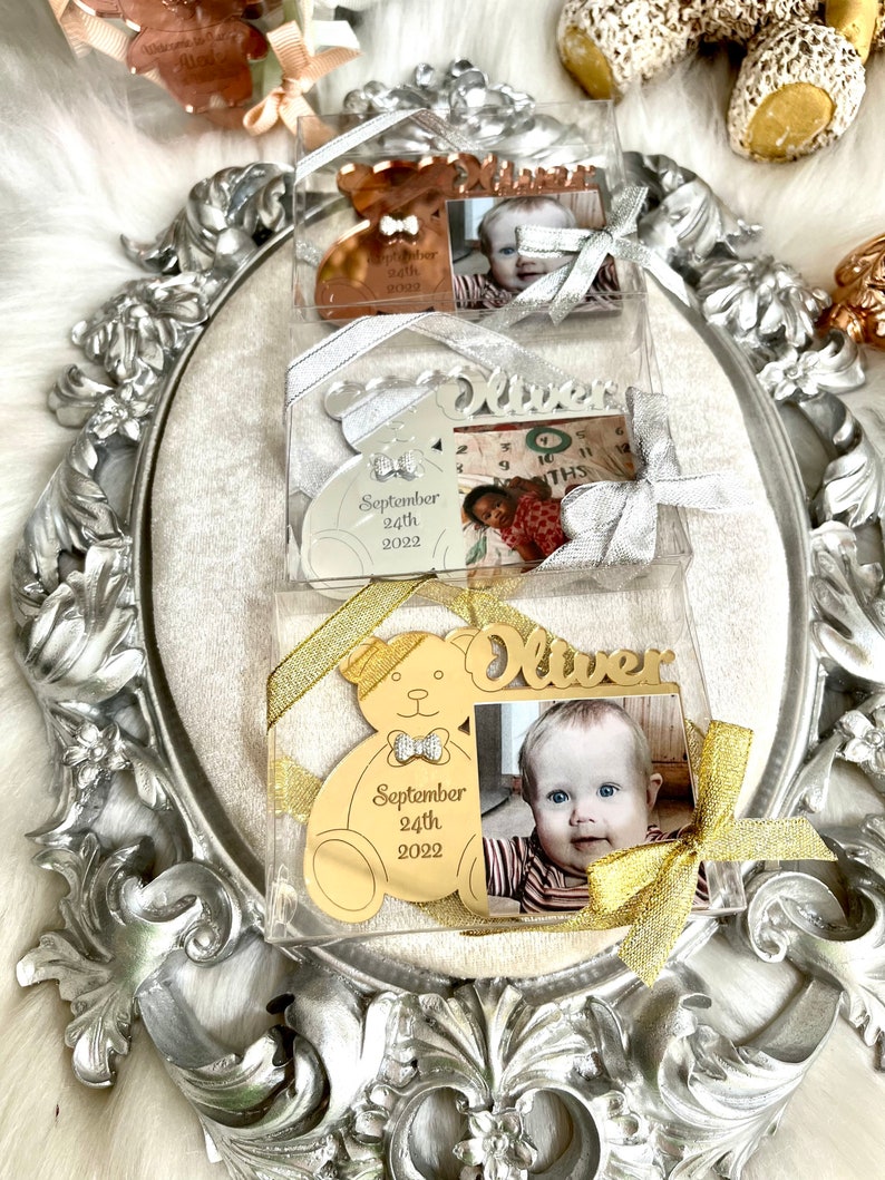 May include: Three gold-colored teddy bear shaped photo frames with a picture of a baby boy. The frames are in clear plastic boxes with gold and silver ribbons. The frames have the baby's name "Oliver" and the date "September 24th, 2022" engraved on them.